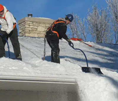 Professional technicians performing roof snow removal with safety harnesses and shovels in Lincoln, NE